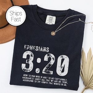 May include: Navy blue t-shirt with the text "EPHESIANS 3:20" in white. The shirt also features a quote from the Bible verse. A gold necklace with a pendant is on the shirt. A brown hat is in the background.