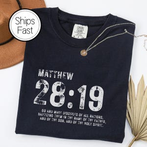 May include: Navy blue t-shirt with the text "MATTHEW 28:19" in white, along with a Bible verse. A gold necklace with a pendant rests on the shirt. A brown hat and a dried palm leaf are in the background.