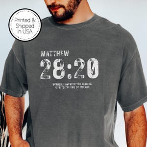 May include: A gray t-shirt with the text "MATTHEW 28:20" in a distressed white font. Below the numbers is the phrase "BEHOLD, I AM WITH YOU ALWAYS, EVEN TO THE END OF THE AGE." A circular graphic reads "Printed & Shipped in USA."