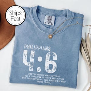 May include: A blue Comfort Colors t-shirt with the text "Philippians 4:6" in white. The shirt also features a gold necklace with a pendant. The text on the shirt reads, "Don't be anxious about anything, but in everything by prayer and petition with thanksgiving let your requests be made known to God."