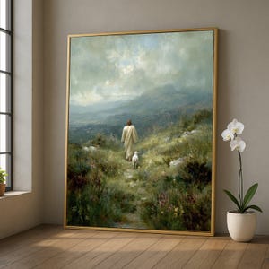 Puede incluir: Pintura de paisaje enmarcada que representa una figura con una túnica blanca caminando con un cordero por un sendero a través de una ladera cubierta de hierba. La obra de arte tiene un marco dorado y un cielo nublado con montañas al fondo.
