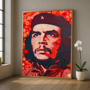 以下が含まれることがあります： ベレー帽をかぶった男性の額装肖像画。赤と黒の色合いで、ドット模様で表現されています。アートワークは金色のフレームで、ニュートラルな壁に展示されています。背景には鉢植えの白い蘭があります。