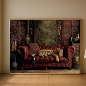 May include: A framed artwork featuring a tiger resting on a red leather sofa. The background includes a landscape painting and ornate wooden panels. The artwork is set in a room with wooden floors and a neutral wall.