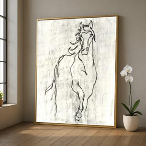 Op de afbeelding: Een ingelijst kunstwerk met een goudkleurige lijst, met een zwarte lijntekening van een paard tegen een getextureerde, gebroken witte achtergrond. Het paard is afgebeeld in een rennende pose, met een minimalistische stijl. Het kunstwerk voegt elegantie toe.