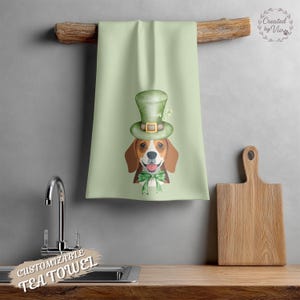 May include: A light green tea towel featuring a beagle wearing a green leprechaun hat and bow tie. The towel is hanging on a wooden rod. A wooden cutting board is on the right. The words "Customizable Tea Towel" are at the bottom.