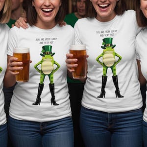 Peut inclure: T-shirts blancs avec un motif de grenouille verte portant un chapeau de leprechaun et des bottes noires. Le texte "YOU ARE VERY LUCKY AND VERY HOT" est imprimé. Les modèles tiennent des verres de bière.