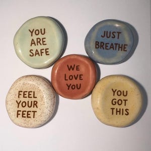 Può includere: Cinque pietre lisce e colorate con messaggi ispiratori. Le pietre sono in tonalità di blu, giallo, arancione e beige. Ogni pietra ha un messaggio diverso in testo marrone: "YOU ARE SAFE", "JUST BREATHE", "WE LOVE YOU", "FEEL YOUR FEET" e "YOU GOT THIS".