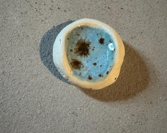 May include: A small, handmade ceramic bowl with a light blue interior and a speckled brown pattern. The exterior is a creamy white color. The bowl has an organic, irregular shape, suggesting it is hand-formed.