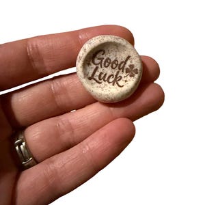 Peut inclure: Un petit jeton en céramique rond de couleur blanc cassé avec une texture mouchetée. La surface présente le texte gravé en brun "Good Luck" accompagné d'une petite icône de trèfle à quatre feuilles et de deux minuscules étoiles.