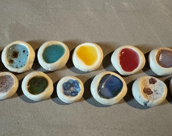 May include: A collection of eleven small, ceramic, pebble-shaped dishes with a variety of colorful, glossy interiors. The colors include blue, green, yellow, red, and purple. The dishes are arranged on a gray surface.
