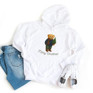 May include: White hooded sweatshirt with a graphic of a teddy bear holding a Christmas tree, wearing a blue jacket and scarf. The text "Merry Christmas" is below the bear. Light blue jeans and white sneakers are also in the image.