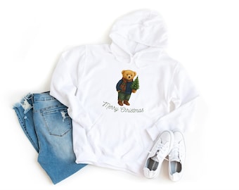 May include: White hoodie featuring a teddy bear graphic in a blue jacket and scarf, holding a Christmas tree, with "Merry Christmas" below. The image also includes light blue jeans and white trainers.