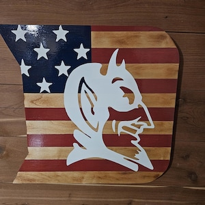 Pode incluir: Uma placa de parede de madeira em forma do estado do Missouri, apresentando um design de bandeira americana pintado com listras vermelhas e de madeira natural, um cantão azul com estrelas brancas e uma silhueta branca de cabeça de diabo.