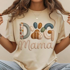 May include: A cream-coloured t-shirt featuring the text "DOG Mama" in a patchwork design. The letters are made of various patterned fabrics, including floral, plaid, and solid colours. A paw print design is in the centre of the word "DOG".
