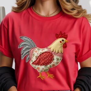 May include: Red t-shirt featuring an appliqued rooster design. The rooster has a cream-colored body, red comb and wattle, and a patterned patchwork design on its chest. The shirt has short sleeves.