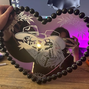 Może przedstawiać: Lustro w kształcie serca z czarną obwódką z koralików. Lustro posiada wytrawiony wzór dwóch szkieletów w kapeluszach kowbojskich, z kwiatowymi akcentami. Lustro jest trzymane przez osobę.