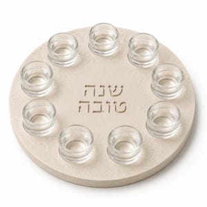 May include: A circular, light beige stone tray featuring ten small, clear glass cups arranged in a ring. The center of the tray is engraved with the Hebrew text "שנה טובה". The stone surface has a textured, matte finish, while the glass cups are smooth and transparent.