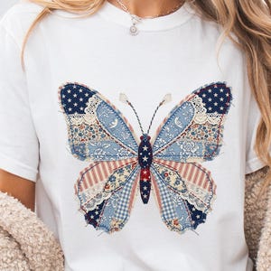 Op de afbeelding: Wit T-shirt met een groot vlinderontwerp. De vleugels van de vlinder zijn een patchwork van blauwe, rode en witte stoffen, waaronder sterren, strepen en bloemmotieven. De vlinder heeft gedetailleerde antennes en een centraal lichaam met rode en witte sterren.