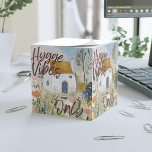 Puede incluir: Un bloc de notas cuadrado con una ilustración en acuarela de una casa de campo con tejado amarillo y puerta azul, rodeada de flores y vegetación. Las palabras "Hygge Vibes Only" están escritas en los laterales con una fuente cursiva. El bloc de notas está sobre una superficie blanca.