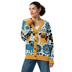 May include: A white cardigan with a mustard yellow trim and black buttons. The sweater features a pattern of pugs, flowers, and abstract shapes in shades of blue, black, and mustard yellow.