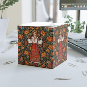 May include: A square notepad with a colourful folk art design. The artwork shows a woman in traditional dress carrying buckets, surrounded by floral and bird motifs in orange, blue, and green. The notepad is on a white surface.