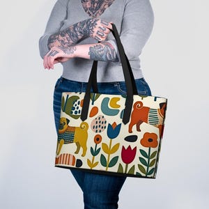 May include: A tote bag featuring a cream background with a repeating pattern of pugs in sweaters, stylized tulips, and abstract shapes in shades of mustard yellow, burnt orange, teal, red, and dark green, finished with black shoulder straps.
