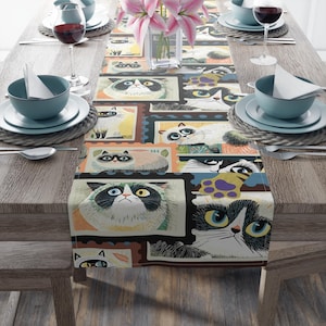 May include: A rectangular table runner featuring a repeating pattern of illustrated cat faces in various colors and poses, resembling postage stamps. The runner is placed on a wooden table set for a meal, with plates, bowls, and wine glasses.
