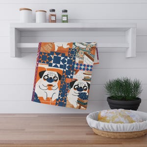 May include: A patterned kitchen towel with a design of pugs, geometric shapes, and kitchen scenes. The towel is predominantly white, with orange, blue, and brown details. It hangs on a white shelf above a wooden surface, with a loaf of bread in a basket.