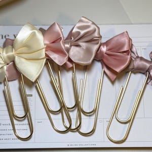 May include: A collection of satin bow paper clips in shades of cream, pink, and mauve. The bows are attached to gold-colored metal paper clips, resting on a white planner page. The planner has text that reads "the y" and "end-of-week assessments".