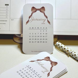 May include: A stack of small, rounded-corner cards featuring monthly calendars for 2020. Each card has a watercolor bow design and the month's name. A gold and white polka-dot pen rests nearby. The Golden Journal Co. is printed at the bottom.