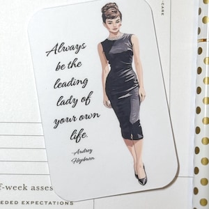 May include: A white card with rounded corners featuring an illustration of Audrey Hepburn in a black dress. The card includes the quote "Always be the leading lady of your own life" and the name "Audrey Hepburn".