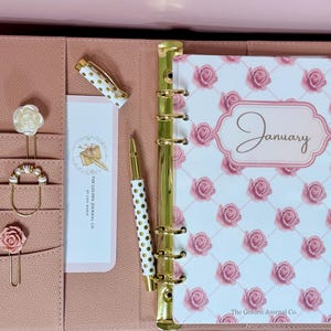 May include: A dusty pink planner with a gold six-ring binder mechanism, featuring a January cover with a pink rose pattern. Includes a white and gold polka dot pen, six decorative rose-themed paper clips, and a card labeled The Golden Journal Co.