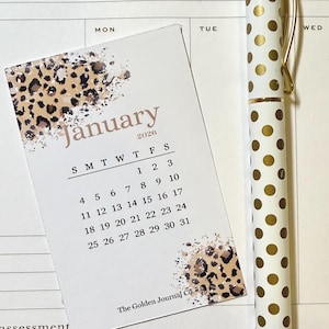 May include: A white pen with gold polka dots and gold accents rests next to a small calendar card. The card displays the month of January 2026, with a leopard print design and the text "The Golden Journal Co."