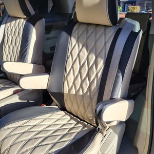 Puede incluir: Fundas de asientos de coche en el interior de un vehículo. Las fundas son de color crema con un patrón de costura de diamantes y detalles en negro y gris. Los reposacabezas y los reposabrazos también están cubiertos con materiales a juego.