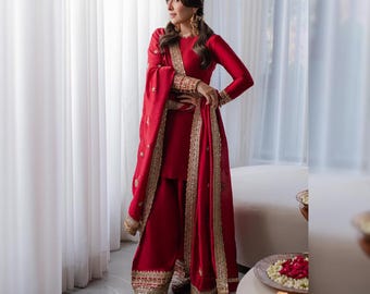 Puede incluir: Un vibrante conjunto rojo que incluye una blusa de manga larga, pantalones de pernera ancha y una bufanda a juego. El atuendo está adornado con bordados dorados en los bordes y puños. La modelo lleva bailarinas rojas y pendientes dorados.