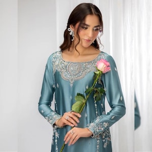 May include: A long-sleeved, dusty blue tunic featuring intricate silver embroidery and beadwork around the neckline and cuffs. The garment is made of a smooth, satin-like fabric and is held alongside a single pink rose with green leaves.