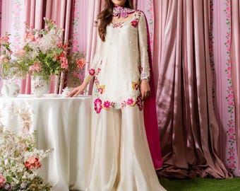 Puede incluir: Un conjunto de color marfil de manga larga con bordados florales en rosa, amarillo y verde. El conjunto incluye una túnica y pantalones de pernera ancha. Una capa rosa y un collar floral completan el conjunto. El fondo presenta cortinas rosas y arreglos florales.