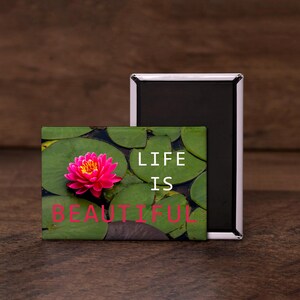 May include: A rectangular magnet featuring a vibrant pink water lily on green lily pads. The words "LIFE IS BEAUTIFUL" are printed in white and pink. A second, black rectangular magnet is behind the first.