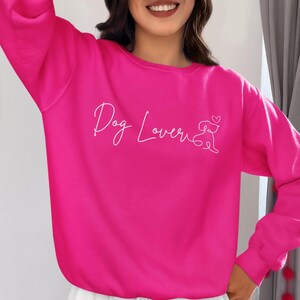 Puede incluir: Una sudadera rosa brillante con las palabras "Dog Lover" en escritura cursiva y una silueta de perro con un corazón. La sudadera es de color liso y tiene mangas largas.
