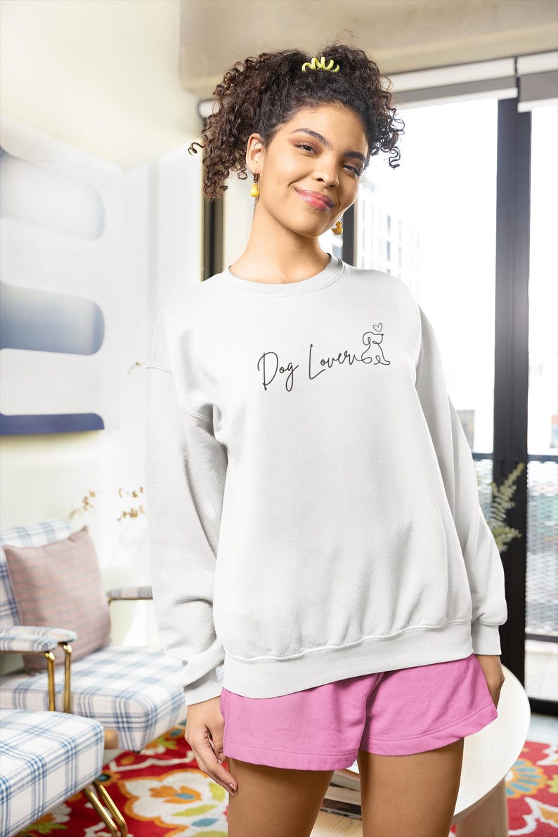 May include: White sweatshirt with the text "Dog Lover" in a script font, along with a small dog silhouette. The person is wearing pink shorts. The background includes a patterned chair and a red rug.