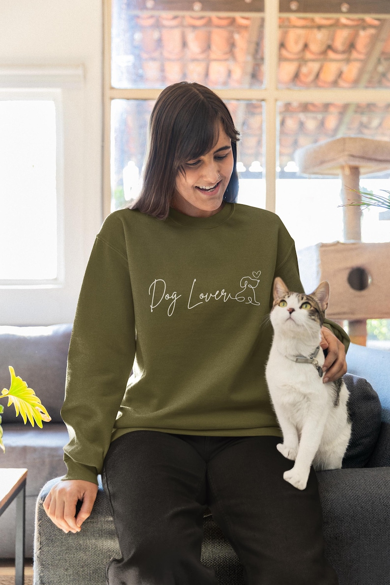 May include: Olive green sweatshirt with the text "Dog Lover" in a cursive font, accompanied by a line drawing of a dog and a heart. The sweatshirt is long-sleeved and made of a soft material.