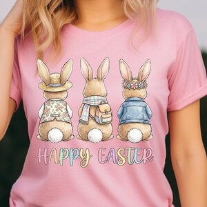 Puede incluir: Camiseta rosa con tres conejos de dibujos animados con diferentes atuendos. Uno lleva sombrero, otro bufanda y el tercero chaqueta. Las palabras "Happy Easter" están escritas en letras coloridas debajo.