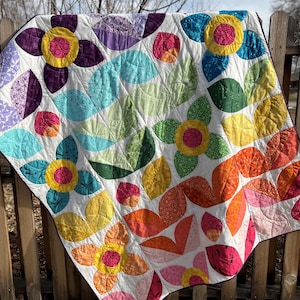 May include: A colorful floral quilt with a white background. The quilt features large, stylized flowers in shades of yellow, pink, orange, blue, and purple, with green leaves. The quilt is draped over a wooden fence.