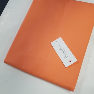 May include: Folded orange fabric with a white tag attached. The tag reads "Baumwolle Fabrics" in a script font. The fabric is a solid color and appears to be cotton or a similar material. The fabric is folded neatly.