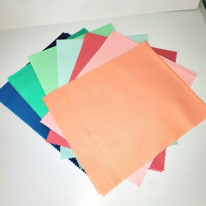 May include: A collection of colorful fabric squares in shades of blue, green, pink, and coral. Each square has a zig-zag edge. The fabrics are arranged in a fan shape, showcasing the variety of colors.