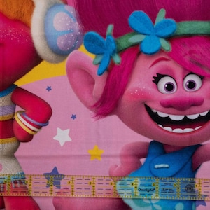 May include: A pink fabric featuring two cartoon characters from the Trolls movie. One character has pink hair and a blue dress, and the other has orange hair and a colorful outfit. The background is pink with stars.