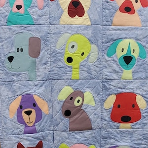 May include: A light blue quilted blanket featuring a patchwork design of various cartoon dog faces. Each dog has a unique color scheme, including shades of blue, yellow, red, and purple. The blanket is likely handmade and suitable for a child's room.