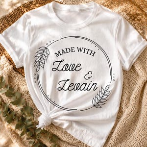 May include: White t-shirt with a round graphic that says "MADE WITH Love & Levain" in black script. The graphic is surrounded by a black circle with leaf accents. The shirt is knotted at the waist.