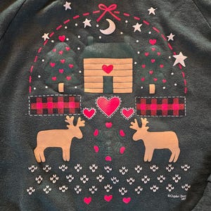 May include: Dark green sweatshirt with a cabin and deer graphic. The design includes a cabin with a heart-shaped door, two deer, hearts, stars, and a crescent moon. The sweatshirt has a crew neck and long sleeves.