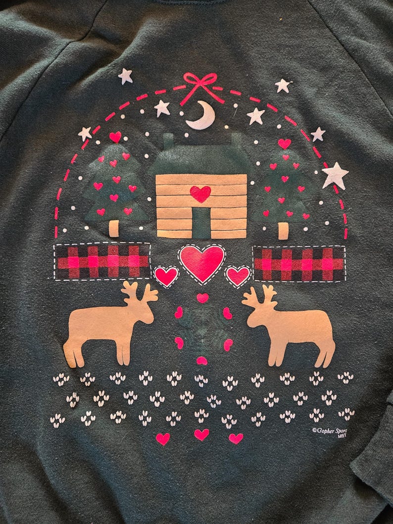 May include: Dark green sweatshirt with a cabin and deer graphic. The design includes a cabin with a heart-shaped door, two deer, hearts, stars, and a crescent moon. The sweatshirt has a crew neck and long sleeves.